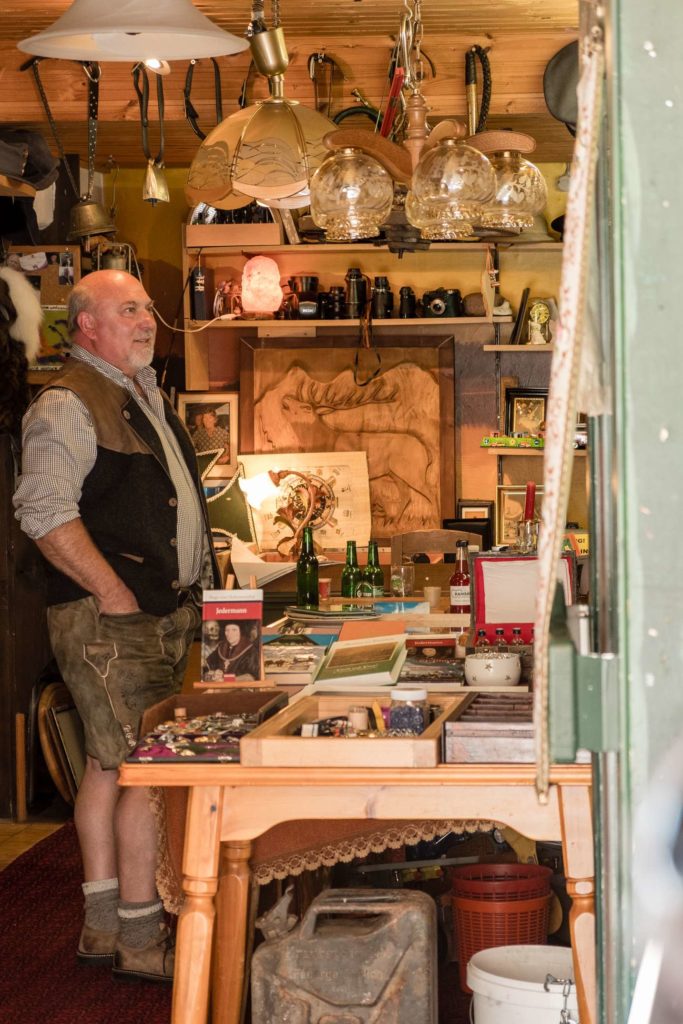 The owner of a secondhand shop in Ramsau am Dachstein stands in this store, dressed in traditional clothes and with a beer in his hand he is talking to a friend or customer outside the photo frame.