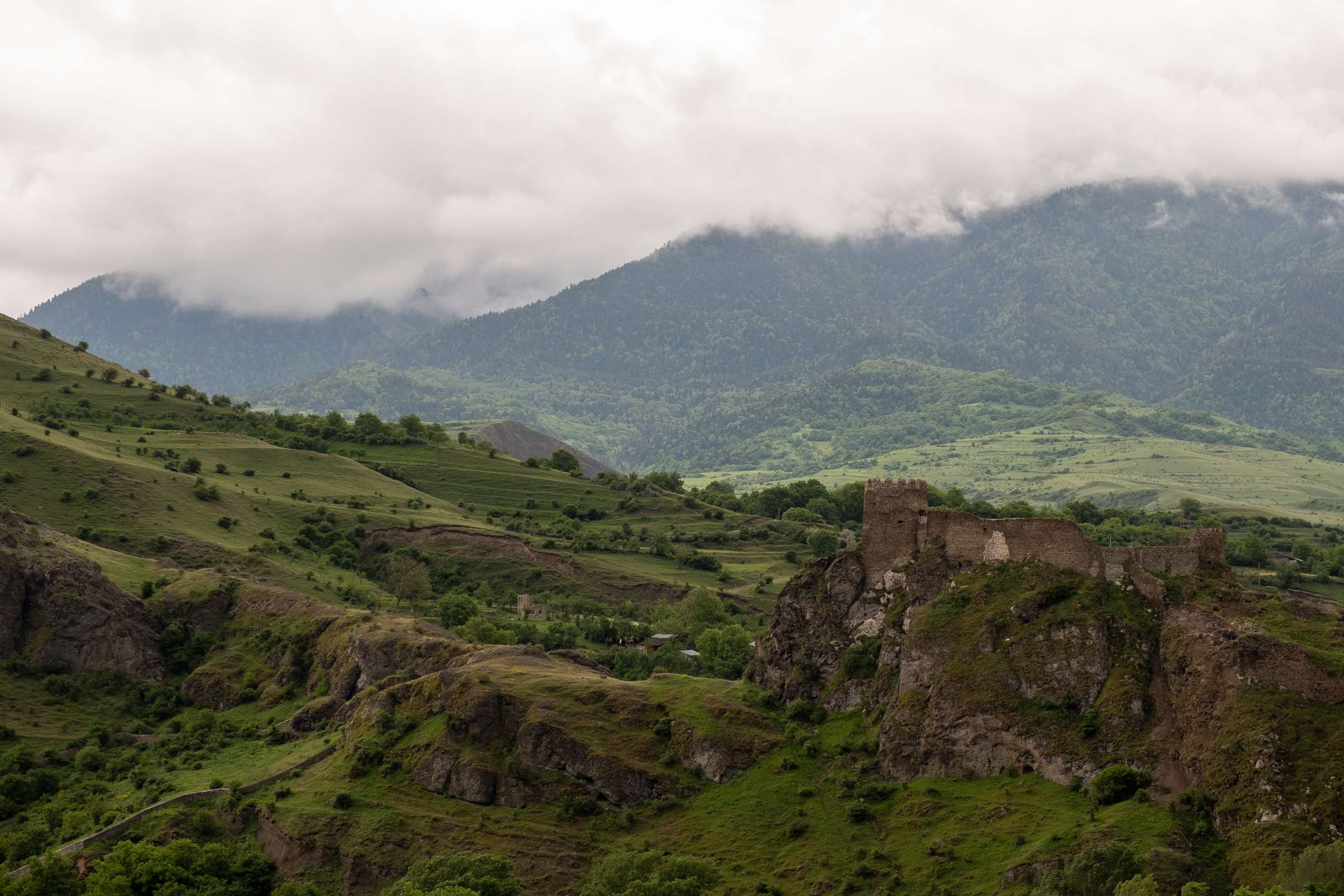 A photo of Atskuri fortress on a cliff between Borjomi and Akhaltsikhe, surrounded by green hills and a cloudy sky