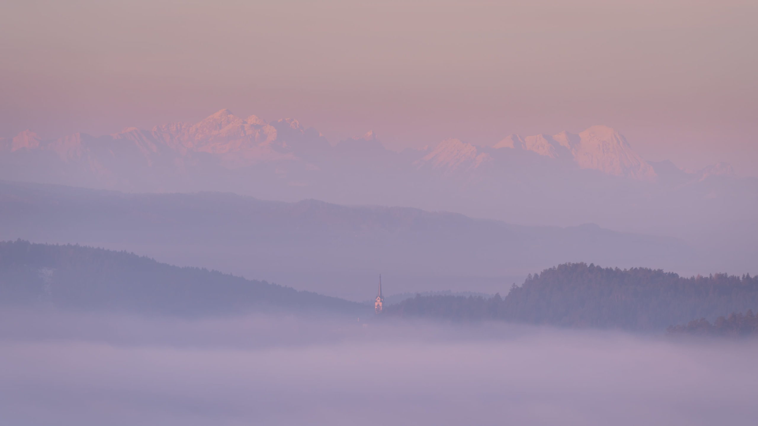 Church spire poking through the thick fog with towering Karawanken mountains behind