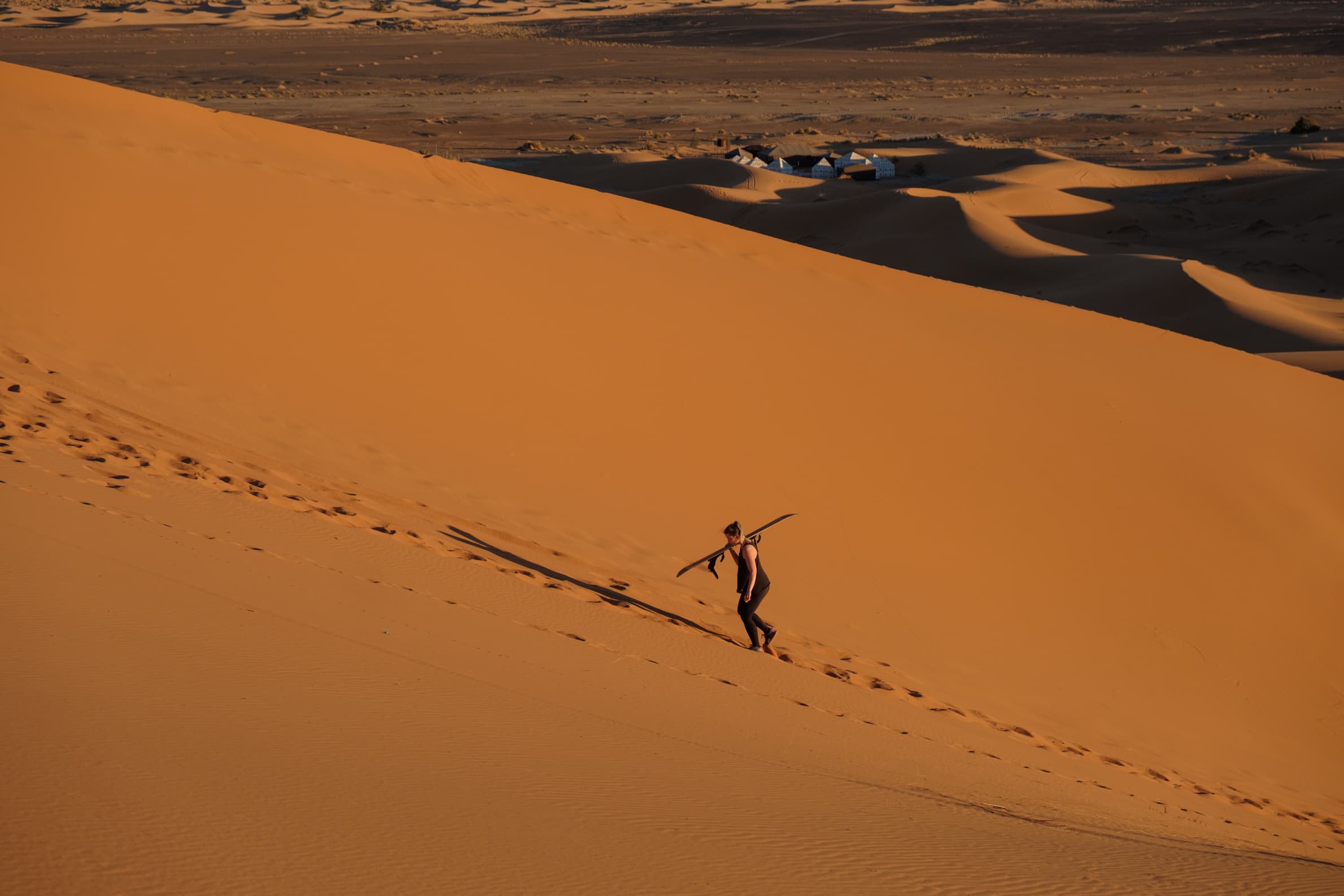 Caroline climbing a sand dune with a snow board with camp in the background