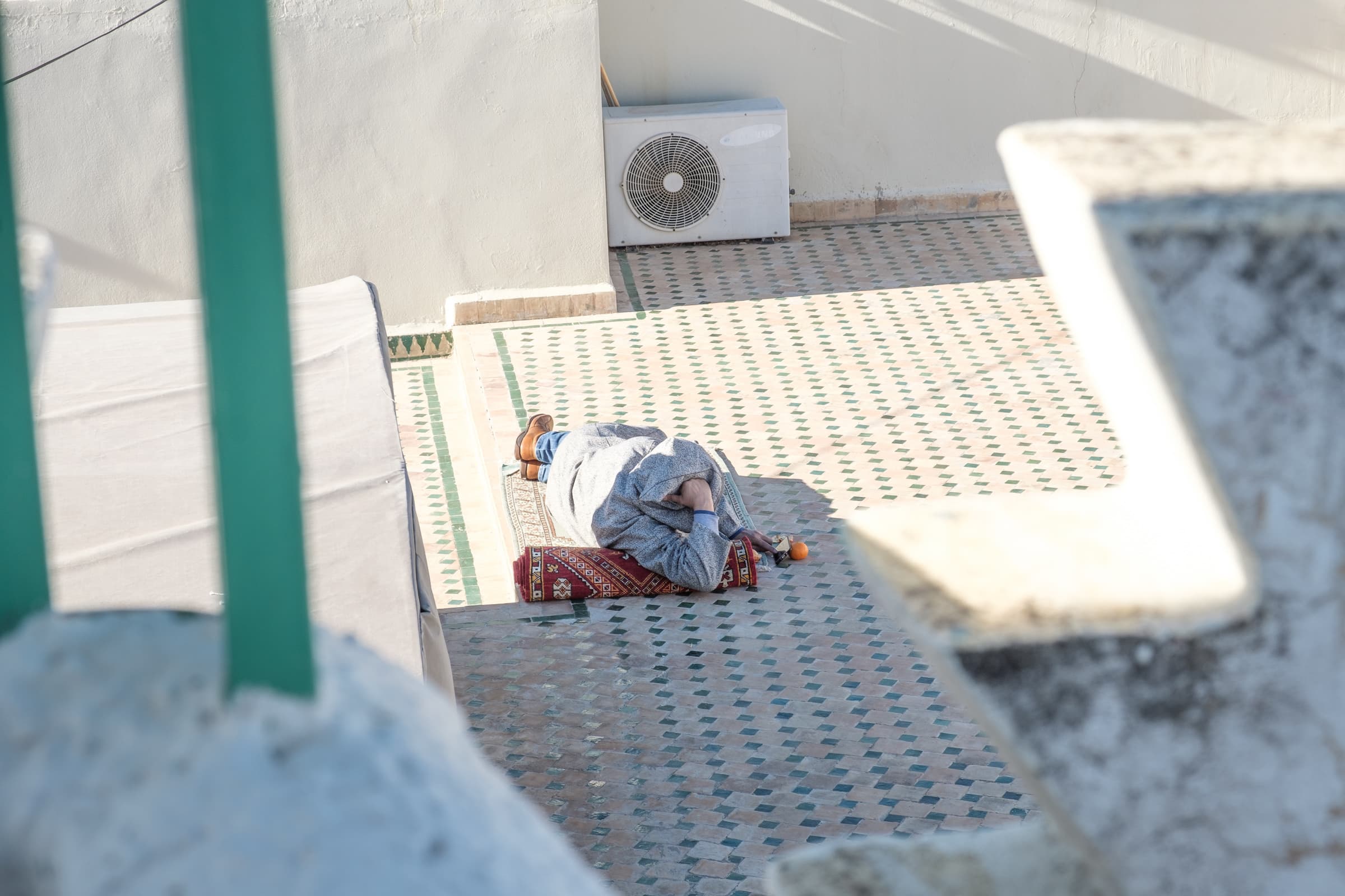 A man lying on a rolled up carpet, watch a video on a iPhone balancing on an Orange, on a rooftop in Fez