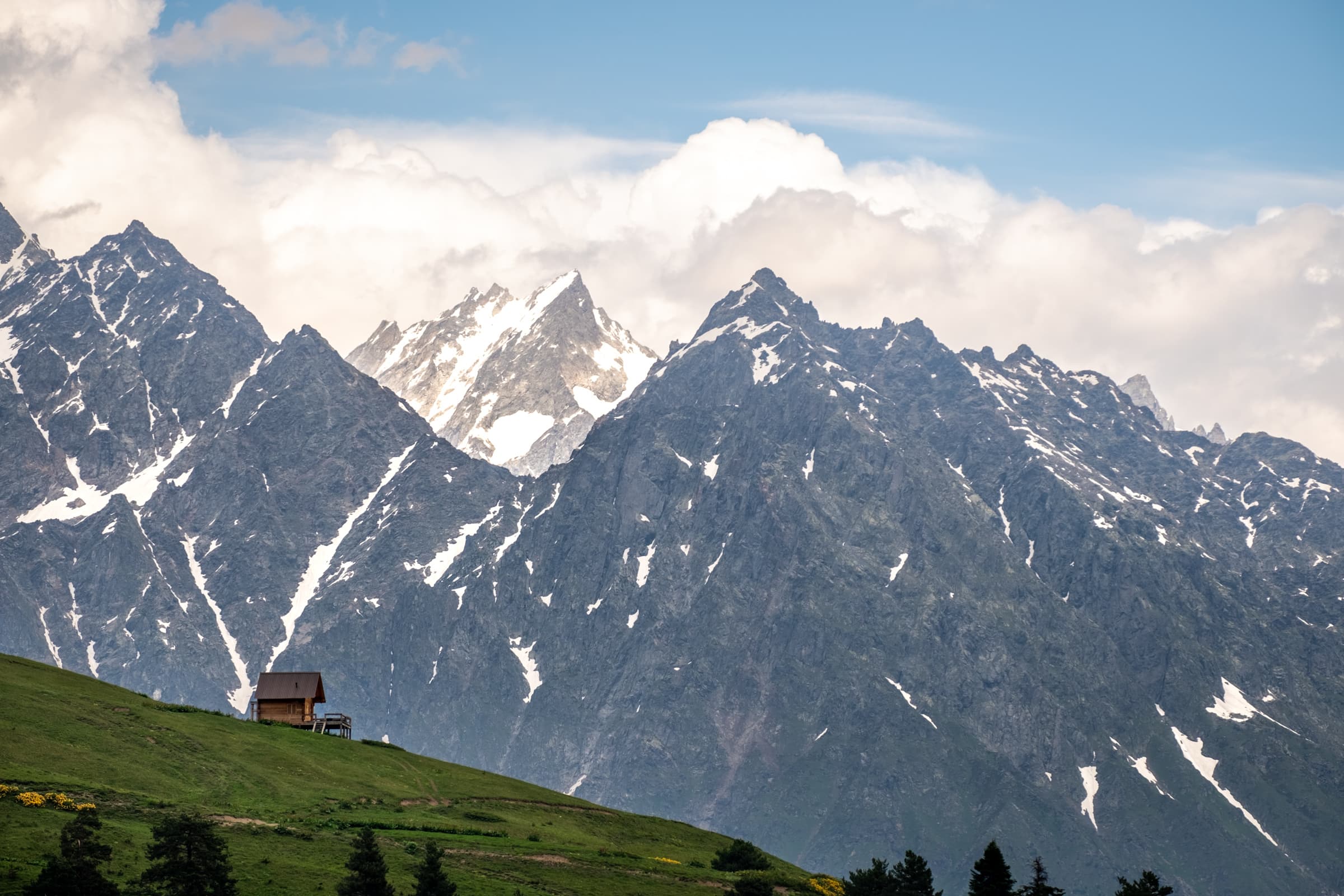 Wooden mountain hut on a green meadow with blue skies and imposing steep jagged mountains behind