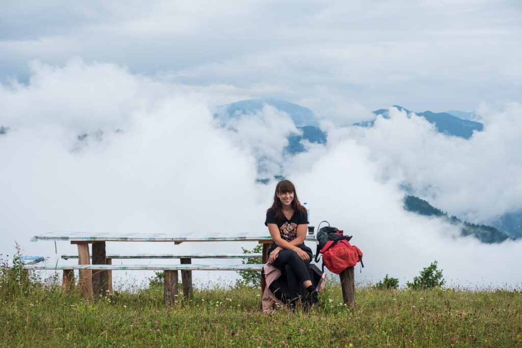 Caroline sitting on a bench smiling with clouds filling the valley and smothering the distant mountains