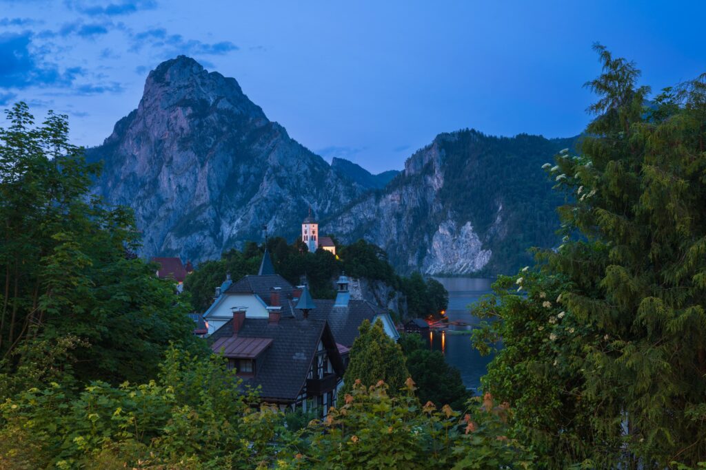 The tiny village of Traunkirchen illuminated at dusk, framed by lush green foliage with Traunstein mountain towering behind