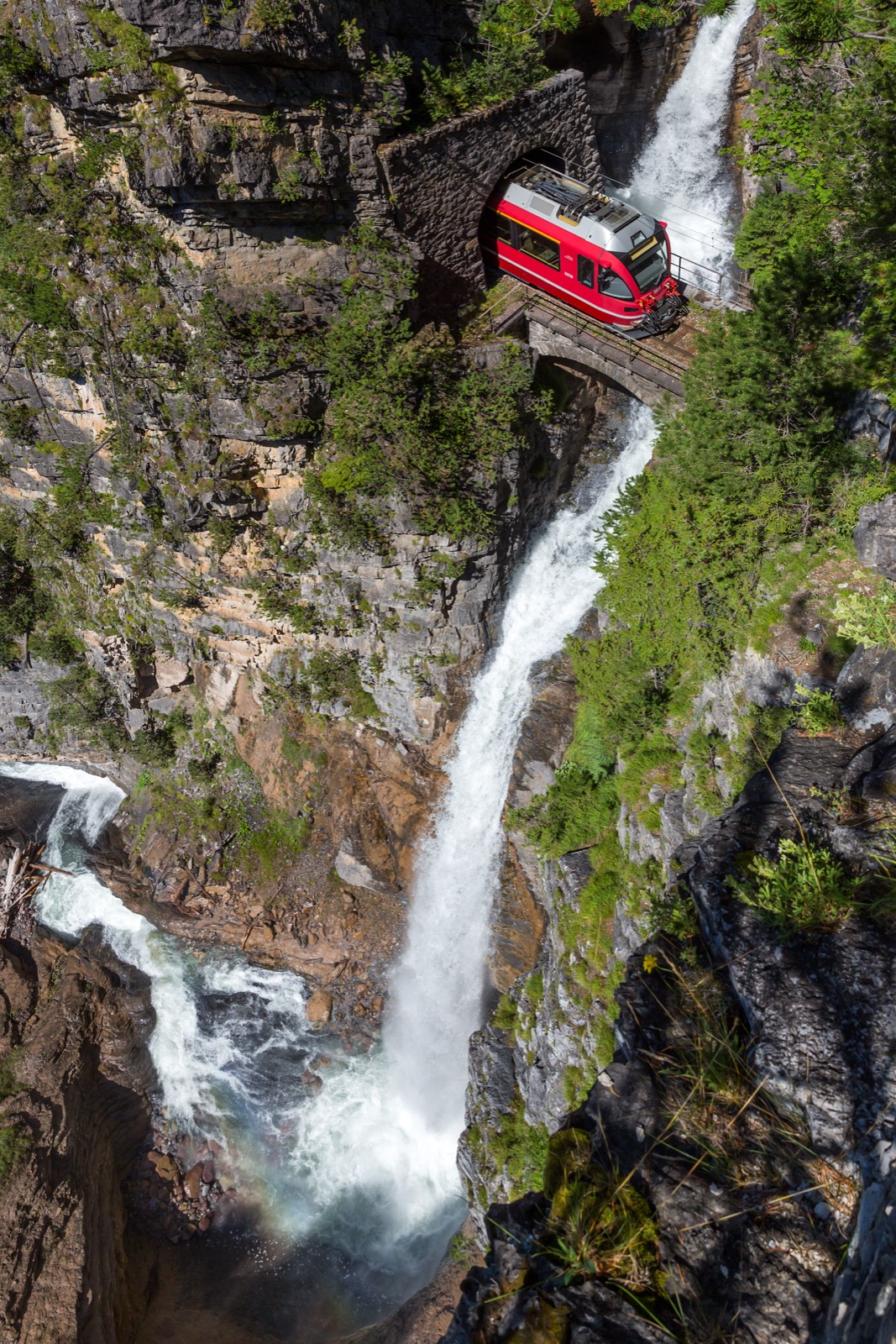 Red Rhaetian Railway train crossing a stone bridge beside a plunging waterfall in a rocky gorge